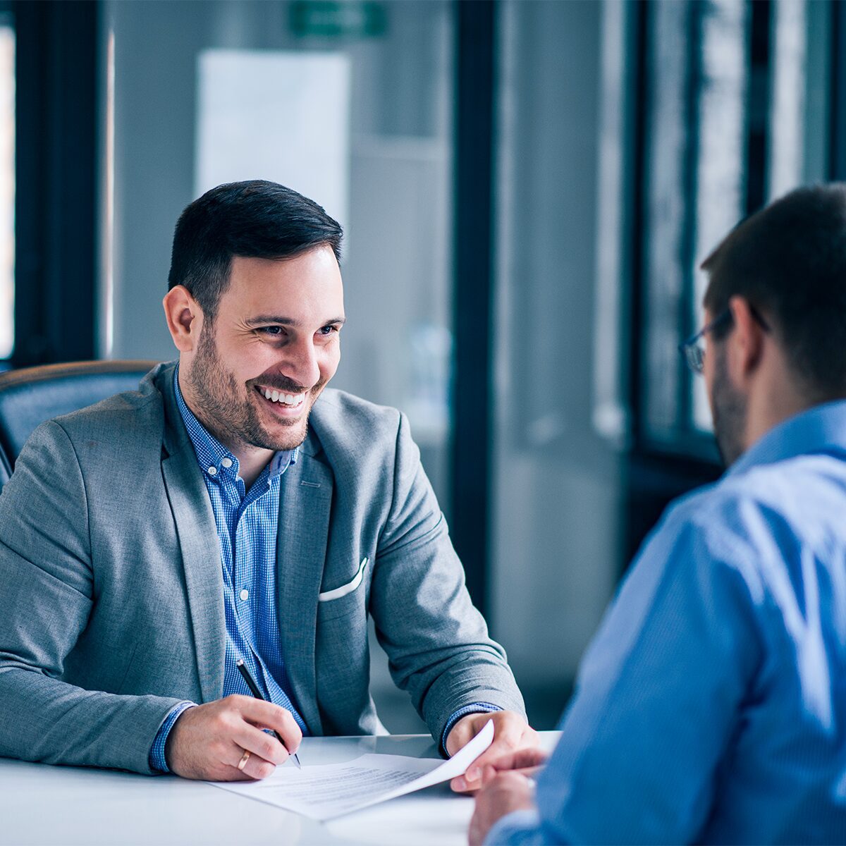 Two men in an office setting, seated at a table. One man, smiling, holds a pen and document, while the other listens attentively.