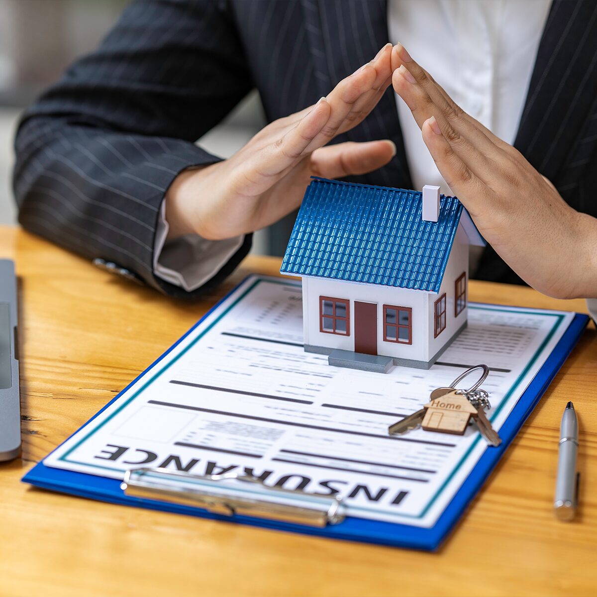 Person in a suit covering a small house model with their hands, sitting at a desk with an insurance document, keys, and a pen.